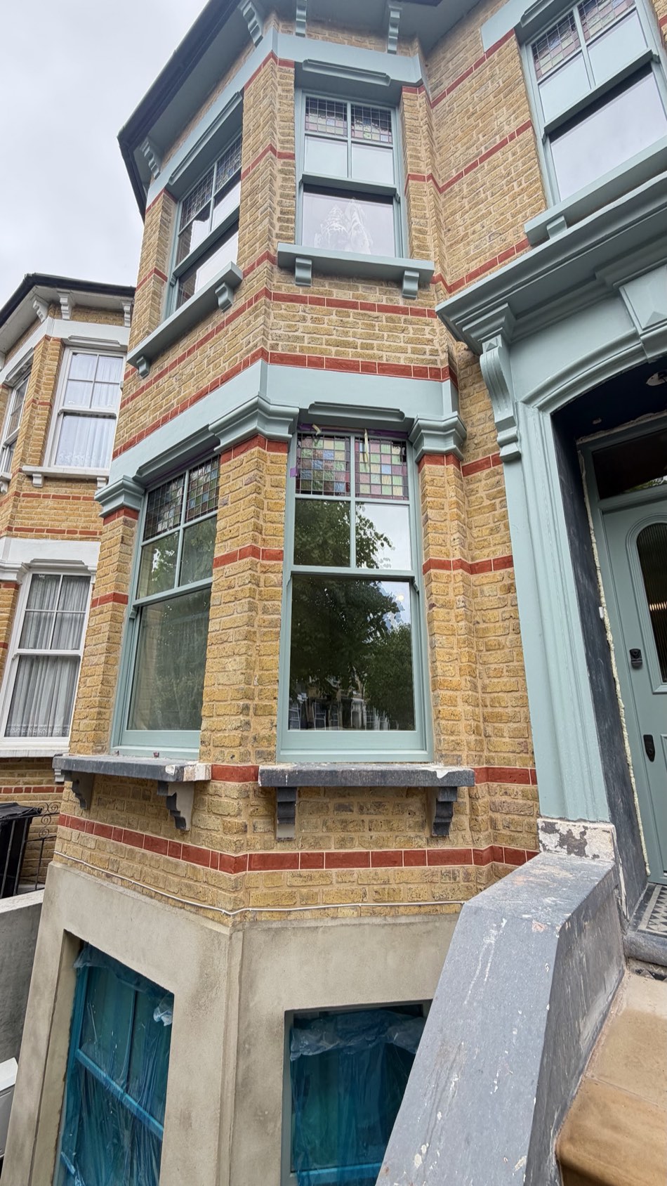 A restored sage-green timber bay window with leaded top lights on a Victorian yellow-brick terrace in East London.