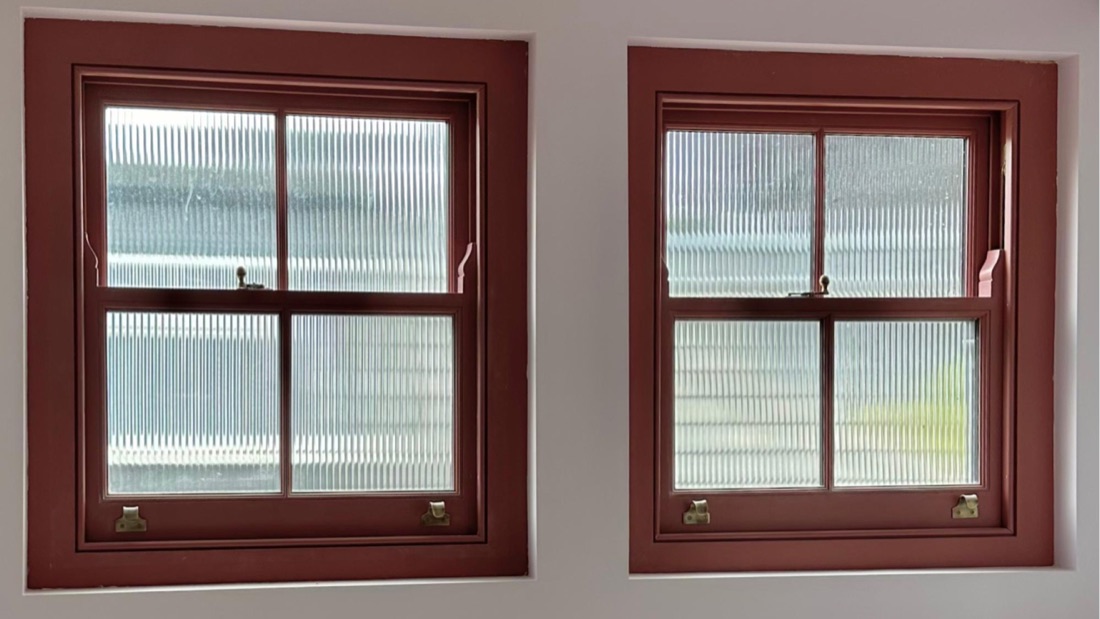 A pair of oxblood-red timber box sash windows with reeded glass and brass sash furniture, fitted into a pale plastered wall.
