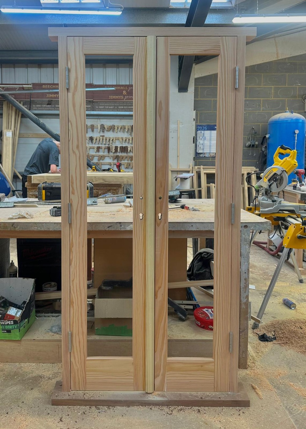 A large arched-panel front door part-built in bare pine on the workbench at Beechhall Joinery's workshop, tools and timber around it.